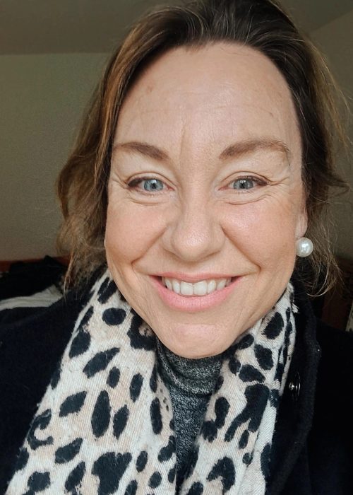 A close-up portrait of Chelsea Banham. She is a woman with light brown hair, blue eyes, and a warm smile, wearing a black coat, a grey turtleneck sweater, a leopard print scarf, and pearl earrings.